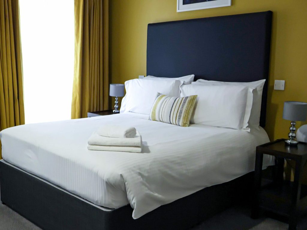 A neatly made bed with white linens, a striped pillow, and folded towels is flanked by two small tables with lamps, set against a mustard yellow wall with a window curtain.