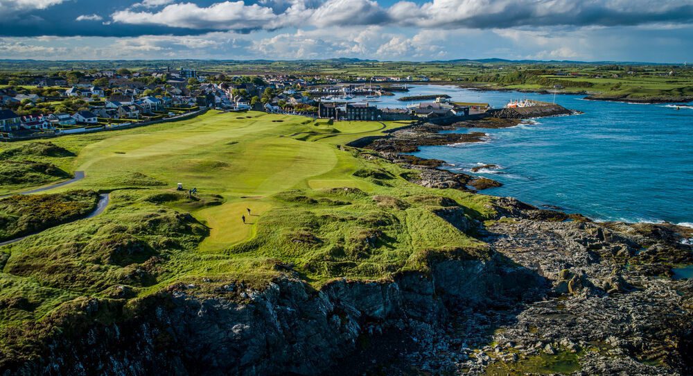 Aerial view of a coastal golf course with rugged cliffs, a small town, and a body of water. The landscape is lush and green under a partly cloudy sky.