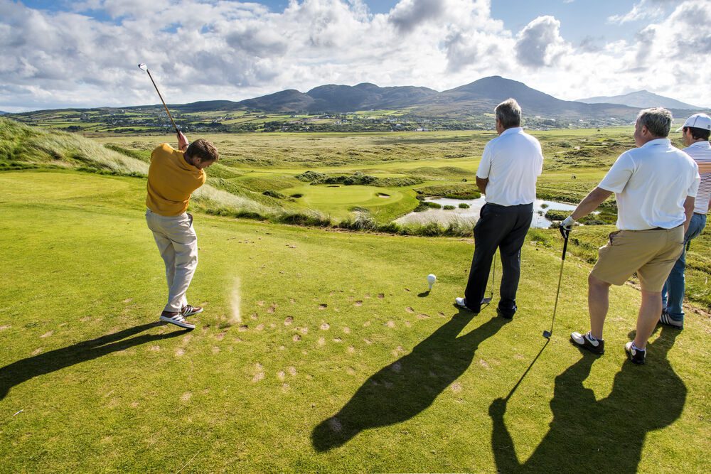 A group of people playing golf on a sunny day, with one person swinging a club. Lush green landscape and mountains in the background.
