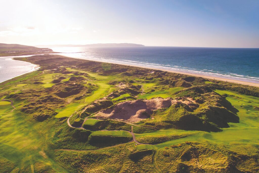 Aerial view of a coastal golf course with rolling green hills, sandy areas, and a beach adjacent to a vast ocean under a clear blue sky.