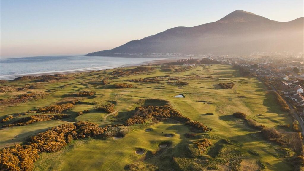Aerial view of a vast golf course by the ocean, with a mountainous backdrop and a town on the right under a clear sky.