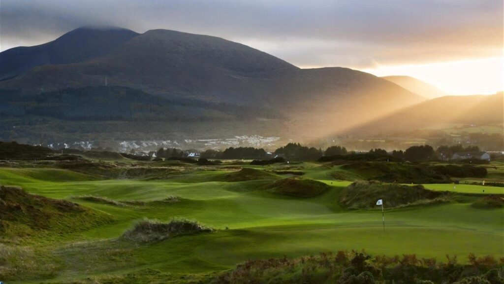 A golf course under sunset light with mountains and a valley in the background. Sun rays illuminate the green landscape, and a flag is visible on the putting green.
