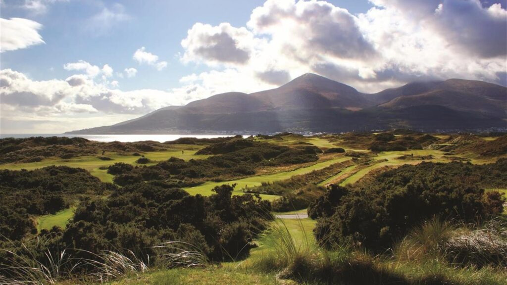 A scenic view of a green golf course with rolling hills and mountains in the background under a partly cloudy sky.