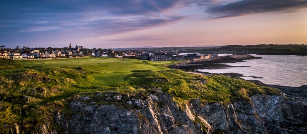 Coastal landscape at sunset with lush green cliffs, a golf course, and a distant town under a cloudy sky.