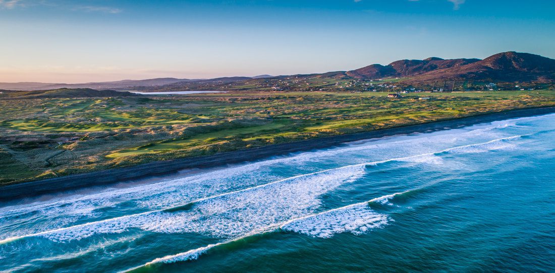 Aerial view of a coastal landscape with waves breaking on a beach, grassy terrain, and distant hills under a clear blue sky.
