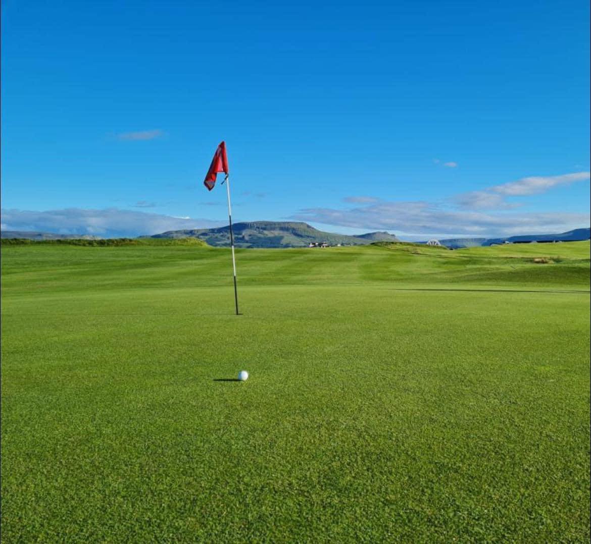 A golf ball rests near the hole on a green course under a clear blue sky with distant hills in the background.