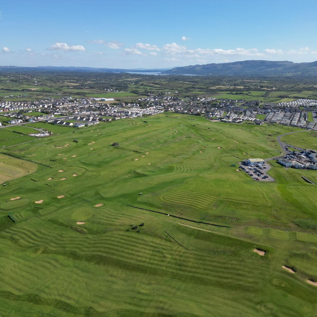 Aerial view of a green golf course with multiple sand bunkers, surrounded by residential areas and hills under a clear blue sky.