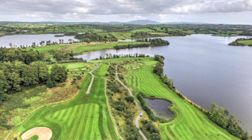 Aerial view of a golf course beside a large body of water, surrounded by lush greenery and hills under a cloudy sky.