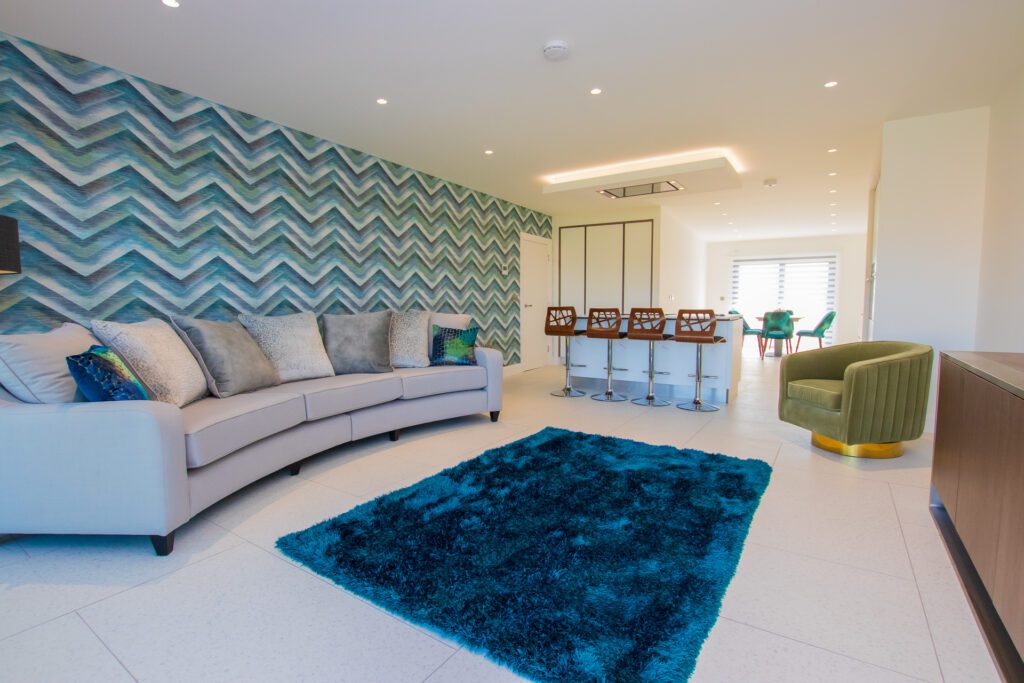 Modern living room with a gray couch, green armchair, zigzag patterned wall, blue rug, and a kitchen island with wooden bar stools.