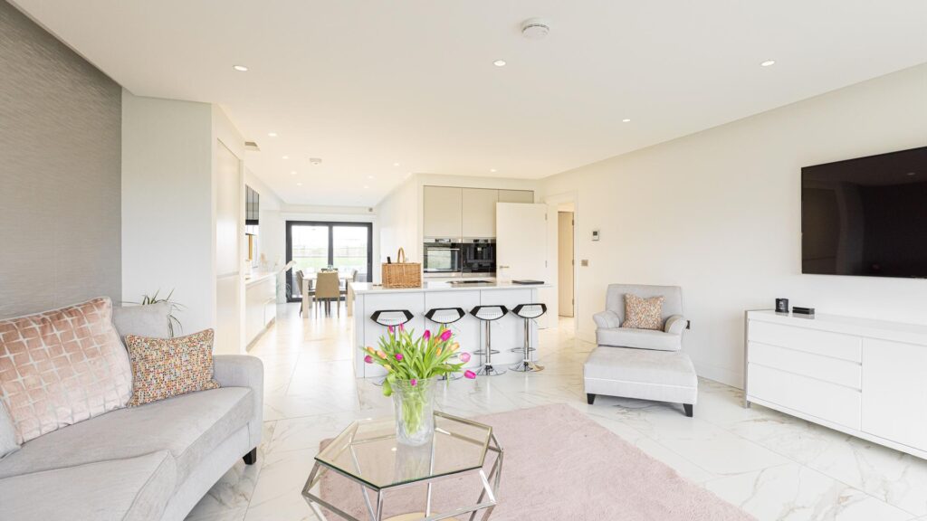 Open-plan living room with beige sofas, a glass coffee table with tulips, and a view into a kitchen with a central island and stools. A dining area is visible in the background.