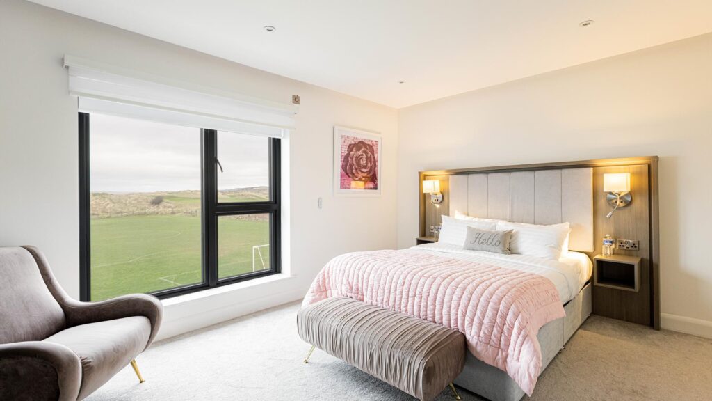 Modern bedroom with a large bed, pink quilt, and a gray armchair. A window shows a grass field outside. A piece of artwork is on the wall above the lamp-adorned nightstands.