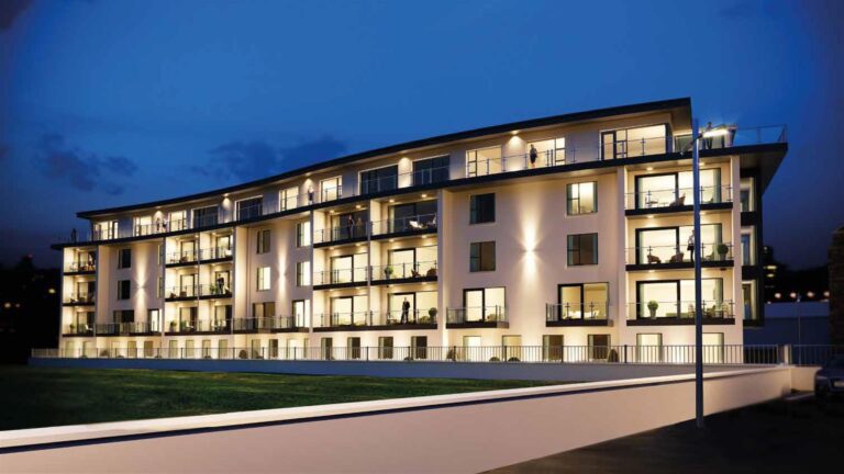 Modern four-story apartment building at dusk, with lit balconies and large windows, alongside a street and grass area.