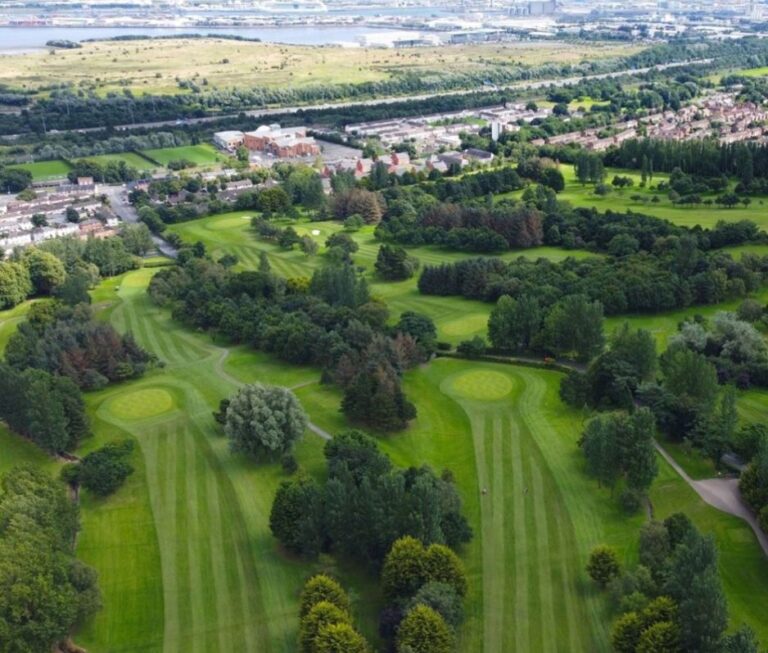 Aerial view of a green golf course with multiple fairways, trees, and a residential area in the background.