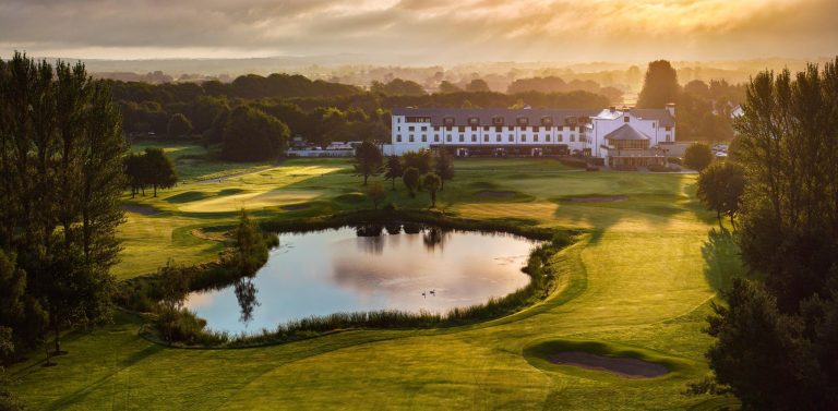 Golf course with a pond, surrounded by trees and a large white building in the background. The sun is setting, casting a warm glow over the landscape.