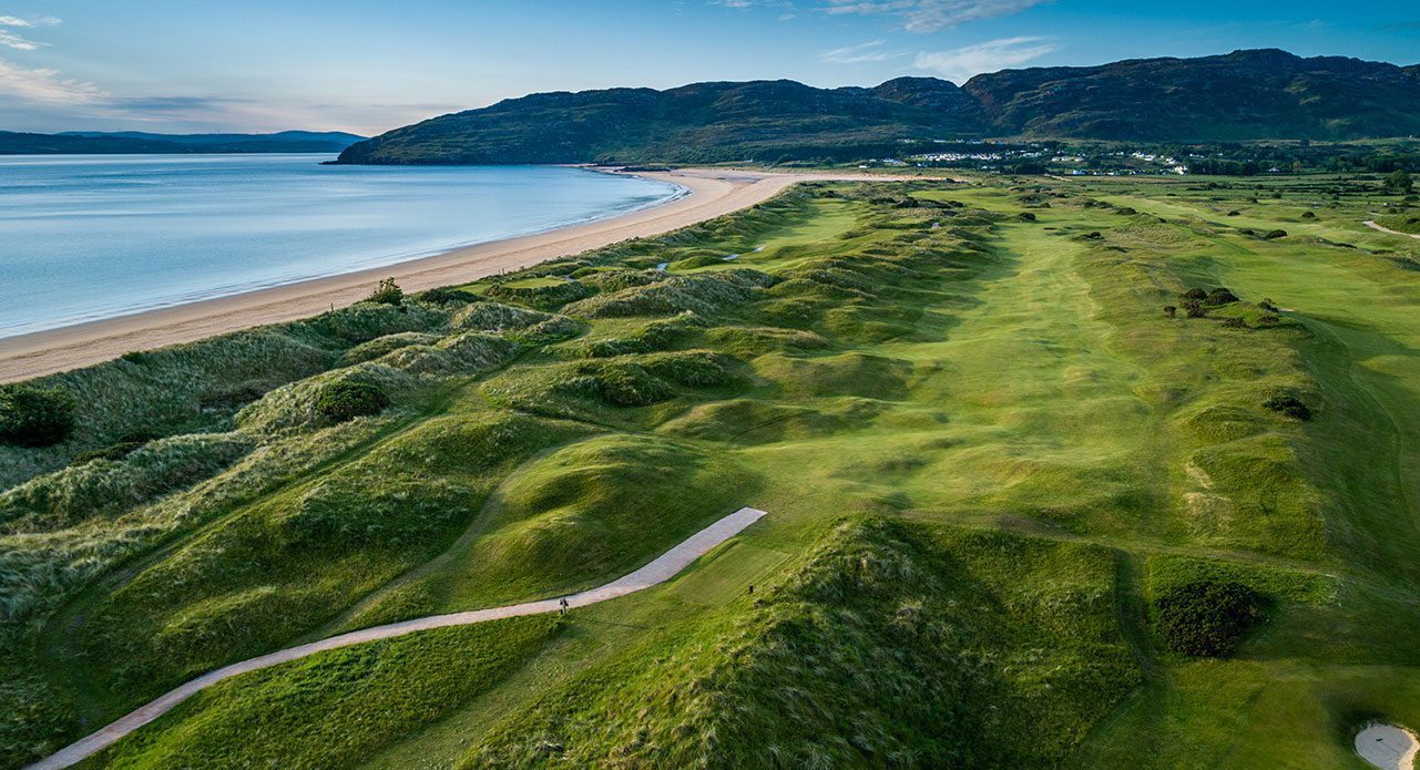 Aerial view of a coastal landscape featuring a sandy beach, grassy dunes, and a winding path, with hills and water in the background under a clear sky.