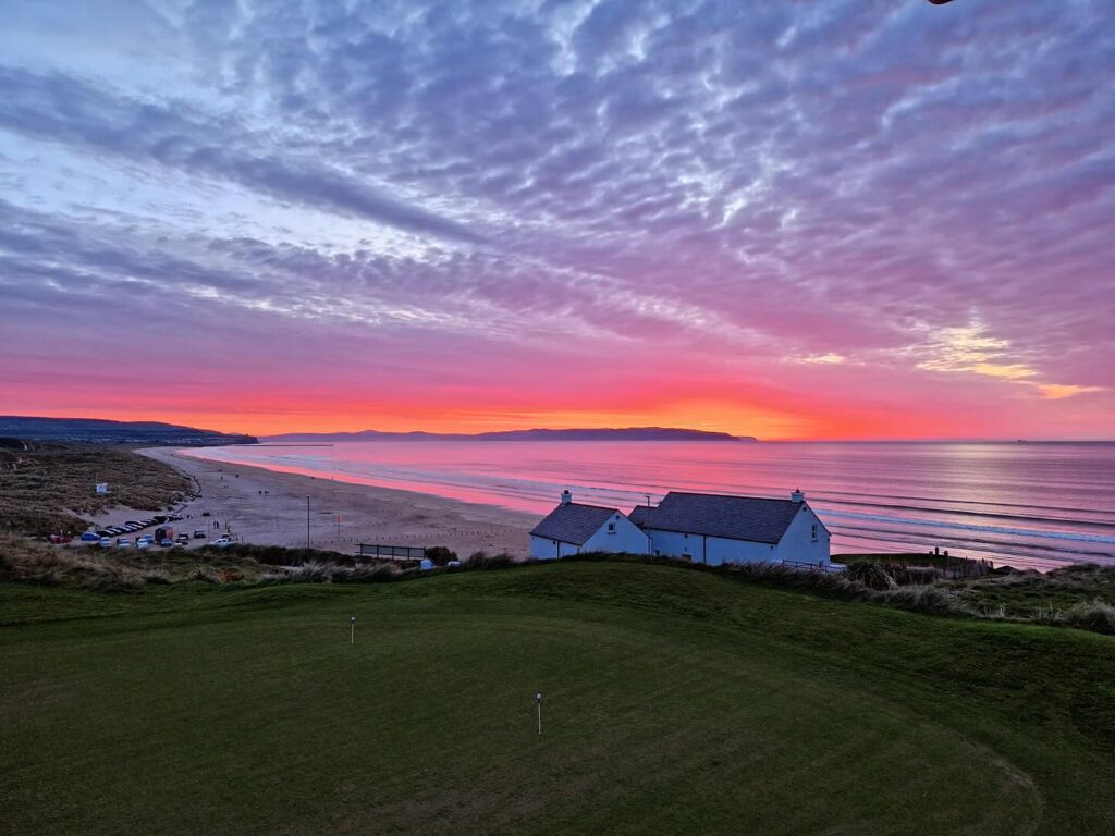 Sunset over a coastal landscape with a white building, grassy area, sandy beach, and ocean waves, under a sky filled with pink and purple clouds.