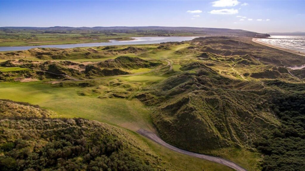 Aerial view of a coastal landscape featuring a golf course with green fairways, surrounding sand dunes, and nearby water. The sky is clear with a few scattered clouds.