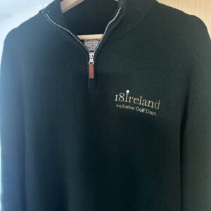 A dark green zip-up sweater with "18ireland Inclusive Golf Days" embroidered in white and gold hangs on a wooden hanger.