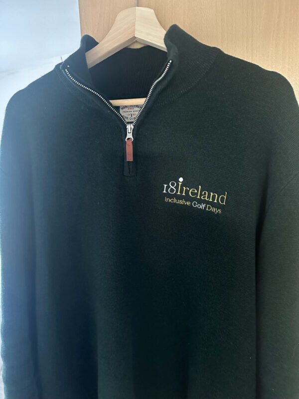 A dark green zip-up sweater with "18ireland Inclusive Golf Days" embroidered in white and gold hangs on a wooden hanger.