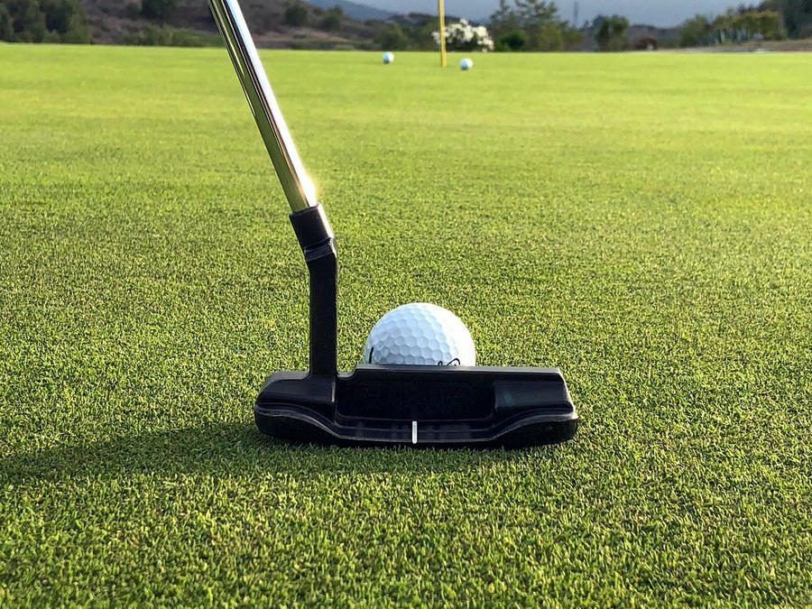 Close-up of a golf ball positioned on a green, ready to be putted with a black putter. In the background, other balls and a flagstick are visible on the golf course.