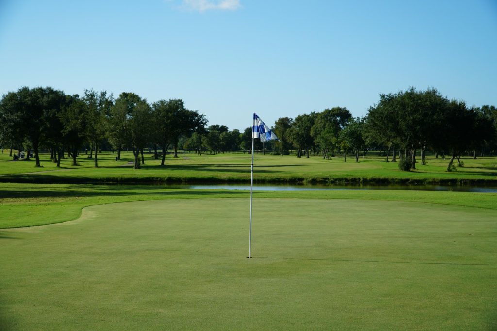 A golf course with a flagstick on the green, surrounded by trees under a clear blue sky.