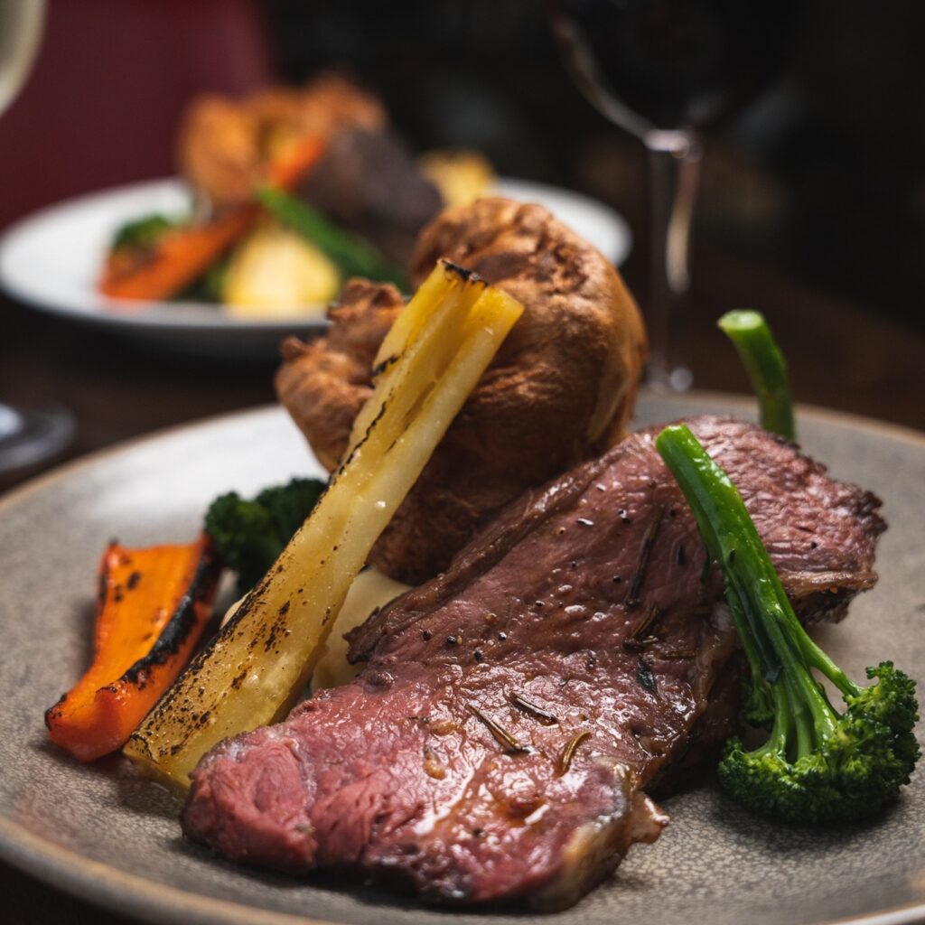 Plate with roasted beef, broccoli, carrots, parsnips, and a Yorkshire pudding. Another similar dish and glasses in the blurred background.
