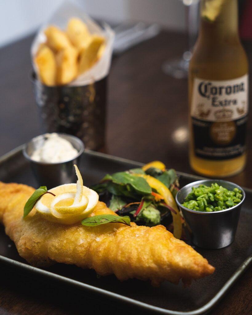 A plate of fried fish with lemon and greens, served with tartar sauce and peas. A bottle of beer and a container of fries are in the background.