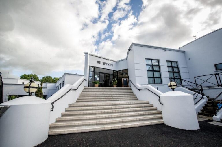 White building with large staircase leading to a reception area. Overcast sky and two round lamps on either side of the steps.