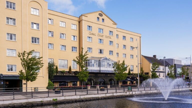 A yellow four-story hotel building labeled "Canal Court Hotel" stands beside a canal with a fountain and tree-lined sidewalk in front.