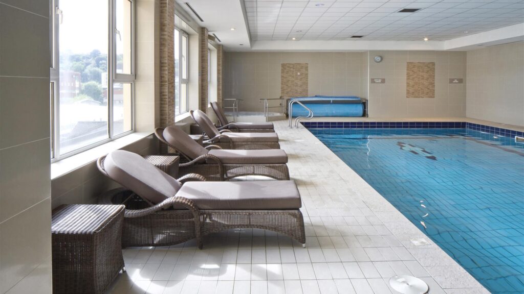 Indoor swimming pool with blue water, wicker lounge chairs, and side tables lined up along a tiled floor beside large windows.