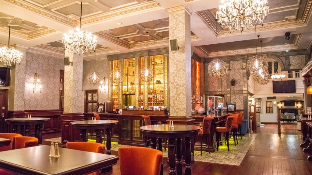 Elegant bar interior with chandeliers, patterned wallpaper, tall wooden tables, orange chairs, and a well-stocked backlit bar counter.