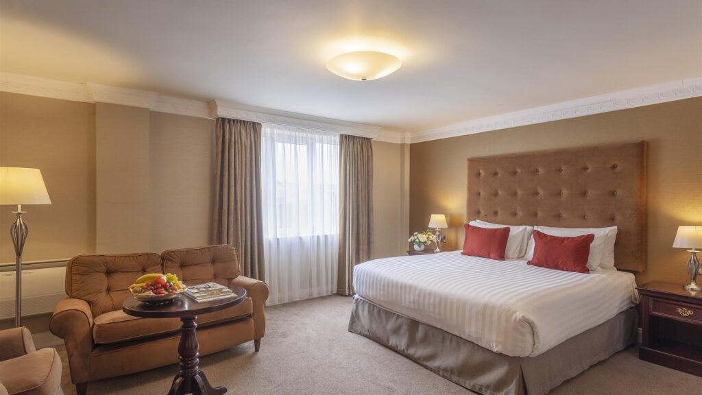 A hotel room with a large bed, two red pillows, a tufted headboard, a sofa, a table with fruit, a nightstand, and a lamp near a window with drapes.