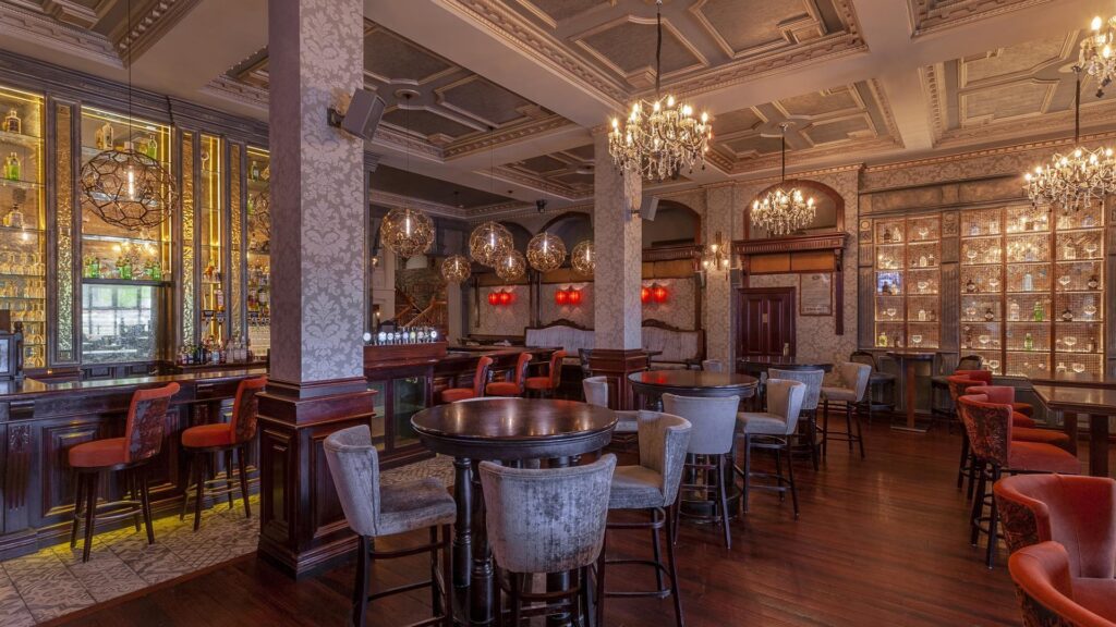 Elegant bar interior with chandeliers, high tables, upholstered chairs, ornate ceilings, and shelves stocked with bottles; warm lighting enhances the classic decor.