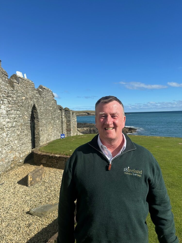 Man in a green sweater stands smiling on a grassy area near stone ruins by the sea under a clear blue sky.