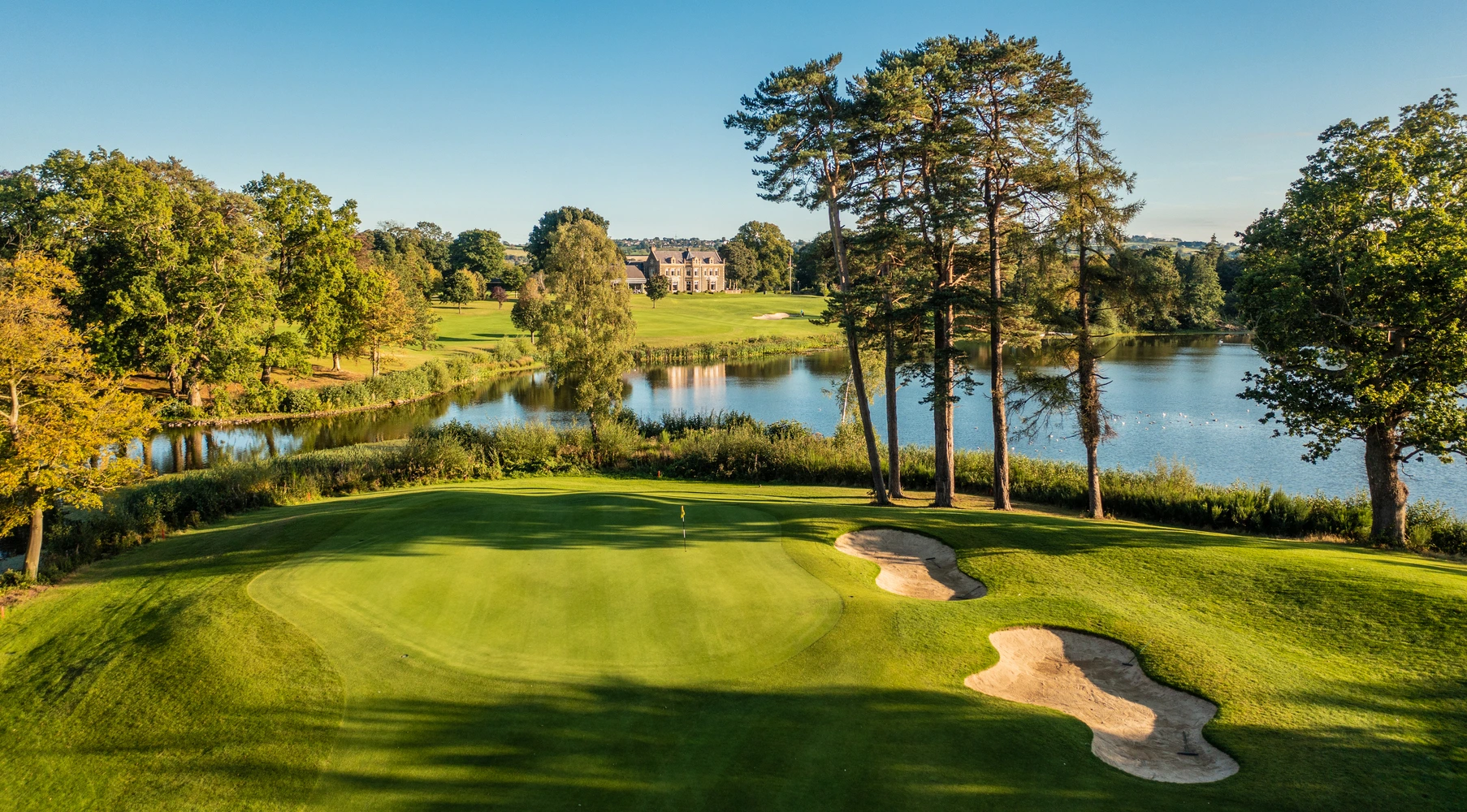 A golf course with a green, sand traps, and trees, adjacent to a lake and a large house in the background under a clear blue sky.