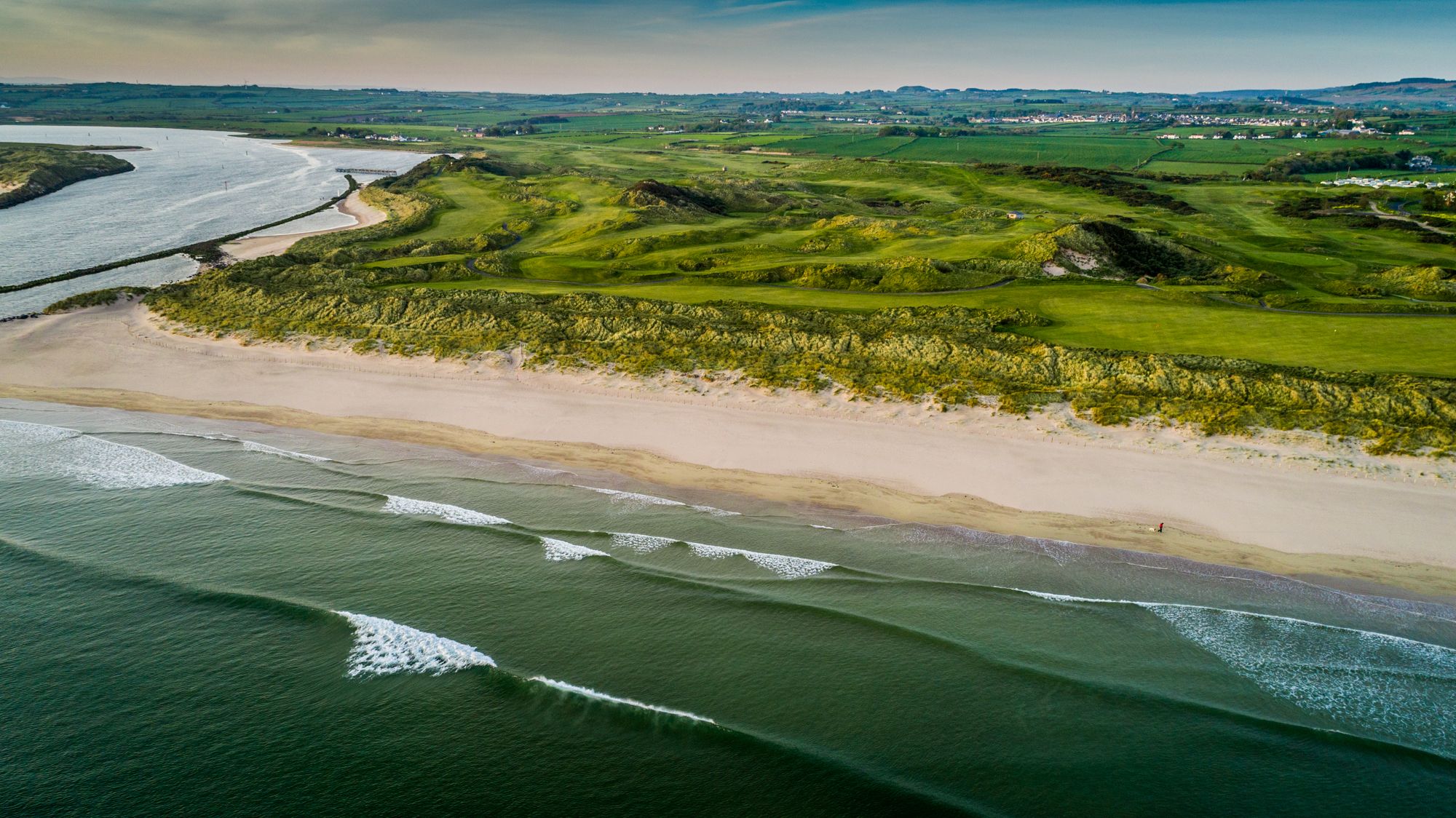 Aerial view of a scenic coastal landscape with a river meeting the sea, green fields, sand dunes, and gentle waves approaching a sandy beach under a clear sky.