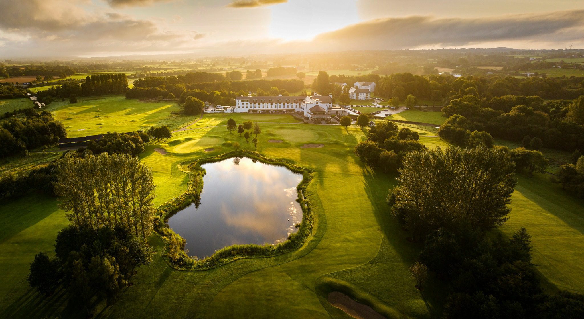 Aerial view of a golf course with a pond, surrounded by trees and greenery at sunset. A white building complex is visible in the background.